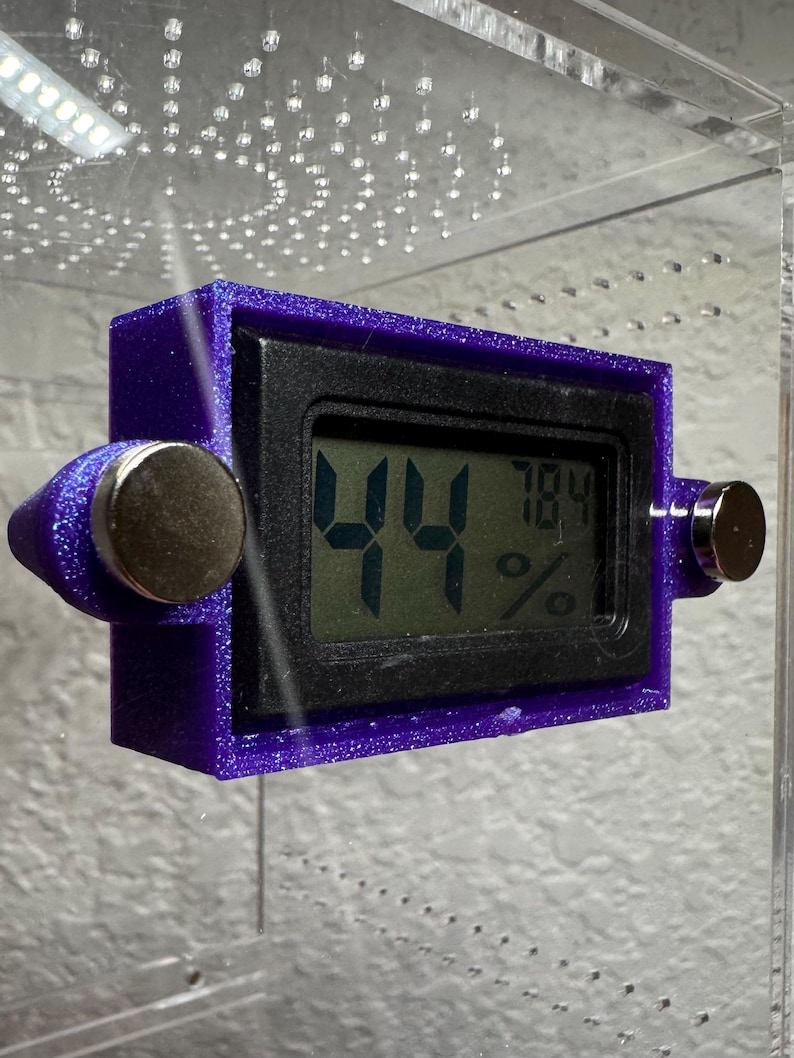 May include: A purple digital hygrometer with a black display showing 44% and 78.4%. The device has two silver magnetic attachments. The hygrometer is mounted inside a clear enclosure.