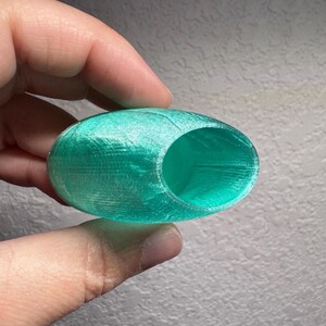 May include: A teal, oval-shaped object with a large circular opening. The object has a textured surface and appears to be 3D-printed. The interior of the object is visible through the opening. The object is held in a hand.