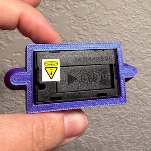May include: A purple rectangular battery holder with a black interior. The holder has a label with a warning symbol and the text "2LR44/1.5V". The holder is being held by a person.