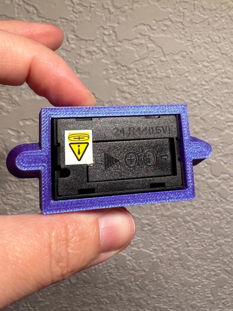 May include: A purple rectangular battery holder with a black interior. The holder has a label with a warning symbol and the text "2LR44/1.5V". The holder is being held by a person.
