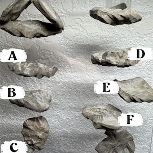 May include: A collection of gray, textured, stone-like shelves and decorative pieces. The items vary in shape and size, including a triangular shelf, a shelf with a glass, and several individual stone-like forms. The pieces are mounted on a textured wall.