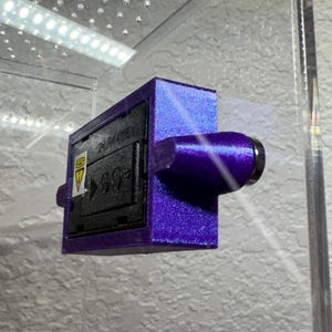May include: A purple rectangular device with a cylindrical extension and a black panel. The black panel has white text and a yellow triangle symbol. The device is mounted inside a clear enclosure.
