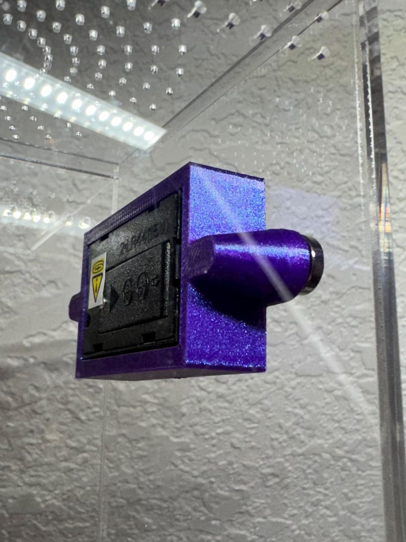 May include: A purple rectangular device with a cylindrical extension and a black panel. The black panel has white text and a yellow triangle symbol. The device is mounted inside a clear enclosure.