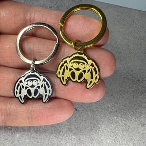 May include: Two keychains with spider charms. One keychain has a silver-colored ring and charm, the other has a gold-colored ring and charm. The spider charms have a detailed design with eyes and legs.