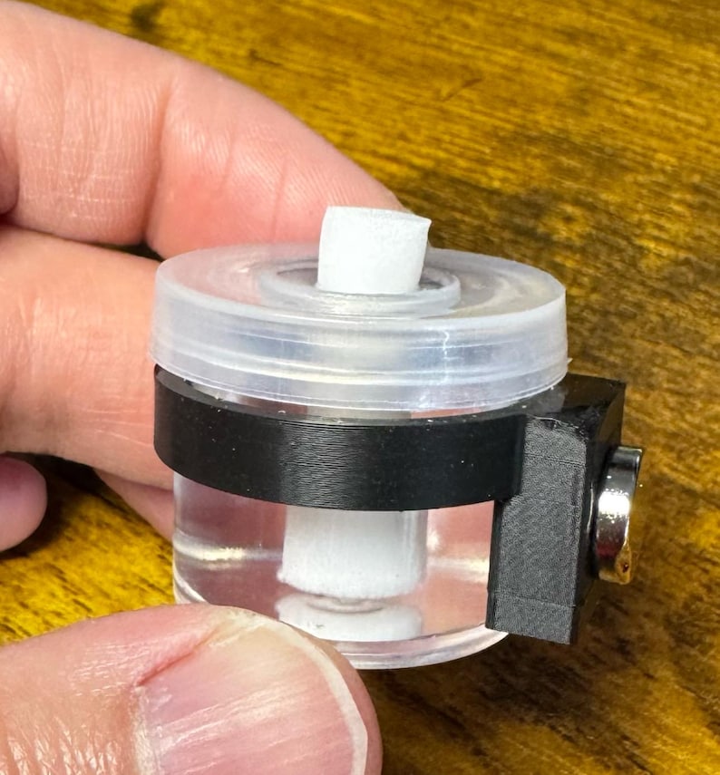 May include: A clear, cylindrical container with a white absorbent material inside. The container has a clear lid and is held in place by a black band and a black rectangular piece with a silver-colored component. The object is held in a hand.