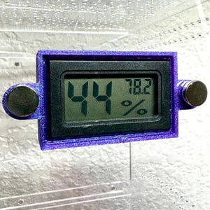 May include: A digital hygrometer with a purple frame displays 44% and 78.2%. The device has two round, black magnets on either side. The screen is black with green digits. The device is likely used to measure humidity and temperature.
