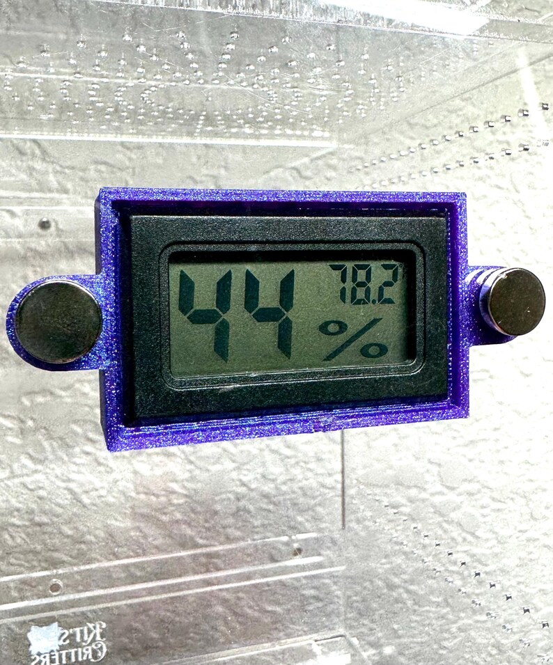May include: A digital hygrometer with a purple frame displays 44% and 78.2%. The device has two round, black magnets on either side. The screen is black with green digits. The device is likely used to measure humidity and temperature.