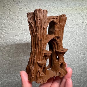 May include: A brown, 3D-printed miniature tree trunk with multiple platforms and openings. The tree has a textured bark appearance and is designed with several small, circular platforms. The overall design suggests a fantasy or nature-themed decorative item.