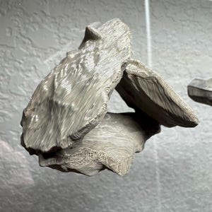 May include: A light gray, abstract sculpture with a textured, layered design. The sculpture has a unique shape with multiple overlapping sections, creating an open space in the center. The surface has a wood-like grain pattern.