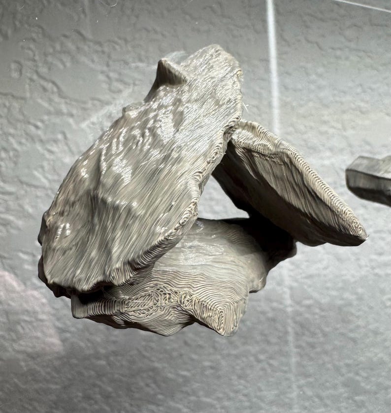 May include: A light gray, abstract sculpture with a textured, layered design. The sculpture has a unique shape with multiple overlapping sections, creating an open space in the center. The surface has a wood-like grain pattern.