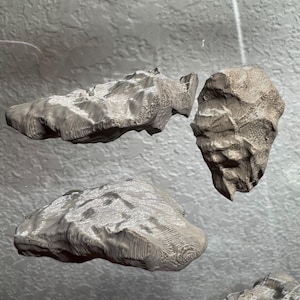 May include: Three gray, textured, rock-like sculptures are suspended in the air. The sculptures have a rough, layered appearance, with visible ridges and patterns. The lighting creates highlights and shadows, emphasizing the three-dimensional forms.