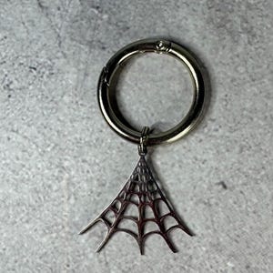 May include: A silver-colored keychain with a circular clasp and a spiderweb charm. The spiderweb charm is triangular and detailed, with a delicate web pattern. The keychain is shown against a gray background.