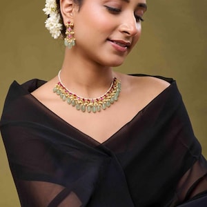 May include: A gold necklace and earring set with green, pink, and gold detailing. The necklace has a series of green, teardrop-shaped stones. The earrings have a similar design. The jewellery is worn with a black off-the-shoulder top.