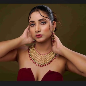 May include: A woman wearing a gold-toned necklace and matching earrings with red and green accents. The necklace has a layered design with a beaded pattern. The woman is wearing a burgundy strapless top.