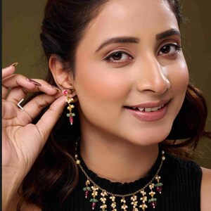 May include: A woman wearing a gold-coloured necklace and earrings set. The jewellery features red, green, and white gemstones. The earrings are being adjusted. The necklace has a cascading design. The woman is wearing a black top.