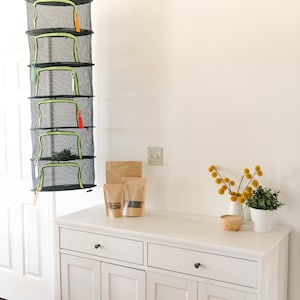 May include: A black mesh herb drying rack with five tiers, each with a green zipper and colorful zipper pulls, hangs from the ceiling. The rack is suspended above a white cabinet with brown paper bags and small plants.