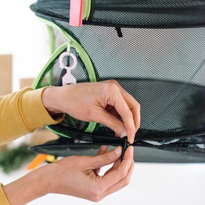 May include: A person is attaching two black clips to a black and green mesh hanging storage unit. The unit has two tiers and is made of a lightweight, breathable material.