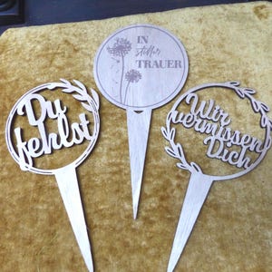 Könnte beinhalten: Drei Gartenstecker aus Holz mit Text. Einer der Stecker trägt die Aufschrift "Du fehlst" in einem kreisförmigen Design mit einem Blätterrand. Ein anderer trägt die Aufschrift "In stiller Trauer" mit einer Pusteblumen-Illustration. Der dritte Stecker trägt die Aufschrift "Wir vermissen Dich" mit einem Blätterrand.