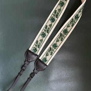 Könnte beinhalten: Ein weißer Kameragurt mit grüner Blumen Stickerei und schwarzen verstellbaren Riemen.