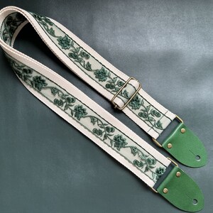 K&ouml;nnte beinhalten: Ein wei&szlig;er Gitarrengurt mit gr&uuml;ner Blumenstickerei und gr&uuml;nen Lederenden. Der Gurt hat eine Messing-Schnalle.