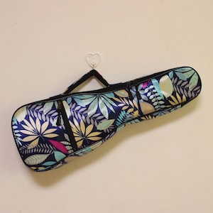 Puede incluir: Una bolsa de ukelele azul con un patrón floral que presenta hojas y flores en tonos azules, verdes, amarillos y rosas. La bolsa tiene dos cremalleras y un asa negra.