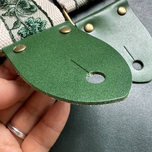 K&ouml;nnte beinhalten: Nahaufnahme eines gr&uuml;nen Ledergitarrengurtes mit zwei Metallschnallen und einem wei&szlig;en Stoffgurt mit gr&uuml;nen gestickten Blumen.