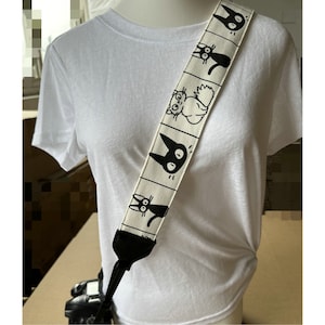 Puede incluir: Una correa de cámara blanca con ilustraciones de gatos negros. La correa es ajustable y tiene una sección de cuero negro en el extremo.