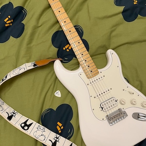 May include: A white electric guitar with a maple neck and a white pickguard, featuring a cream-colored guitar strap decorated with black line-art illustrations of various cats. A single white guitar pick rests near the guitar body.