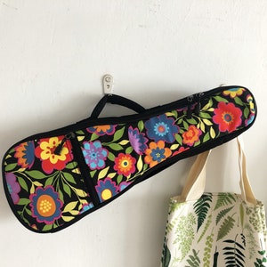 Puede incluir: Una funda negra para ukelele con un vibrante estampado floral en rojo, naranja, amarillo, azul y morado. La funda tiene una cremallera y un asa negras. Una bolsa de mano con estampado de helechos cuelga cerca.