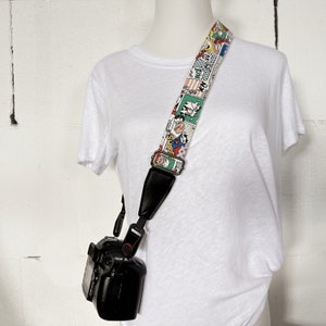 Puede incluir: Una camiseta blanca con una correa de cámara negra con un estampado de dibujos animados coloridos. La correa tiene una hebilla negra ajustable y un accesorio de cámara negro.