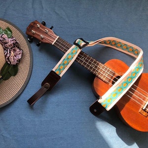 Op de afbeelding: Een ukulele met een ananas-patroonriem. De ukulele is bruin met een lichte houten hals en ligt op een blauwe stoffen ondergrond. Een strohoed met een bloemversiering is ook op de afbeelding te zien.