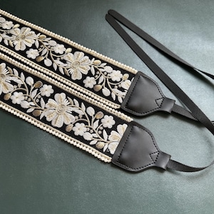 May include: A black camera strap with white and gold floral embroidery and a pearl trim. The strap has two black leather adjustable ends.
