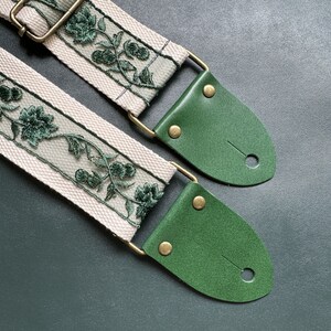 K&ouml;nnte beinhalten: Ein Gitarrengurt mit einer wei&szlig;en Canvas-Basis und einem gr&uuml;nen, gestickten Blumenmuster. Der Gurt hat zwei gr&uuml;ne Lederenden mit Messing-Schnallen.