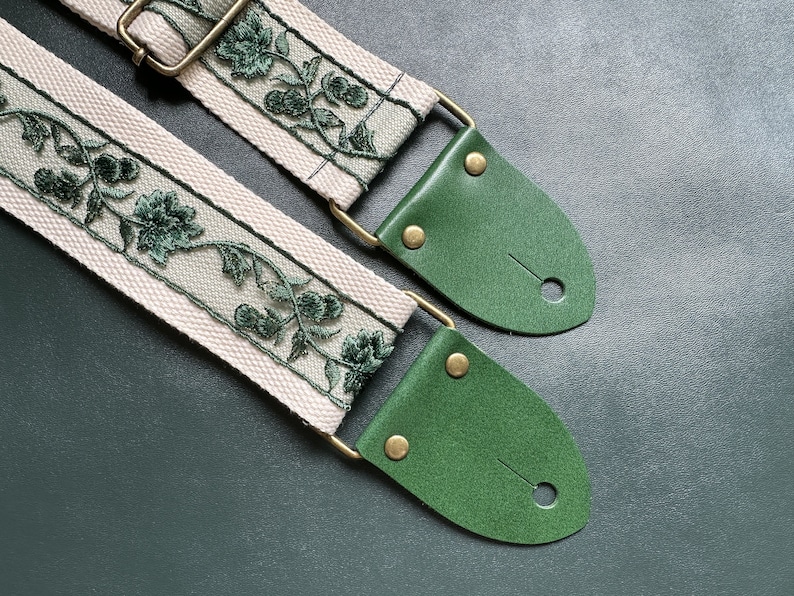 K&ouml;nnte beinhalten: Ein Gitarrengurt mit einer wei&szlig;en Canvas-Basis und einem gr&uuml;nen, gestickten Blumenmuster. Der Gurt hat zwei gr&uuml;ne Lederenden mit Messing-Schnallen.