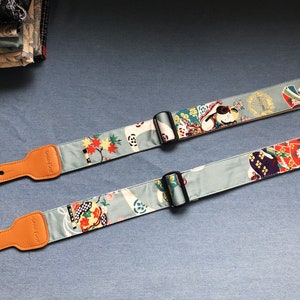 May include: Two guitar straps with adjustable buckles. The straps are made of a light blue fabric with a colourful Japanese-inspired pattern. The straps have brown leather ends.