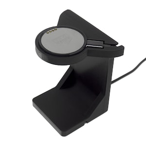 以下が含まれることがあります： 黒い時計充電スタンド。円形の充電パッド付き。パッドはTAG Heuerのロゴが入ったグレーの表面です。スタンドは角度がついており、電源用のケーブルが内蔵されています。