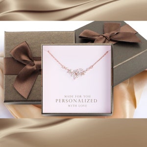 May include: A delicate rose gold necklace with a sparkling cluster of cubic zirconia stones in a floral design. The necklace is displayed in a brown gift box with a brown satin ribbon. The box has a white label with the text "Made for you Personalized with love".