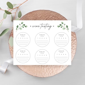 May include: A white wine tasting card with six circles for rating, each with a 1-5 scale. The card is decorated with green leaf accents and the words "wine tasting." The card rests on a copper-colored plate with a white ribbon and greenery.