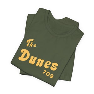 Könnte beinhalten: Olivgrünes T-Shirt mit dem Text "The Dunes 709" in gelber Retro-Schrift. Das T-Shirt ist gefaltet, wobei das Design auf der Vorderseite zu sehen ist. Das T-Shirt besteht aus einem weichen Material.