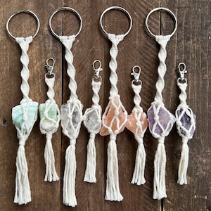 May include: A collection of macrame keychains with various raw crystal stones. Each keychain features a natural stone in a woven net, a silver-tone clasp, and a large metal ring. The crystals are in shades of green, clear, orange, and purple.