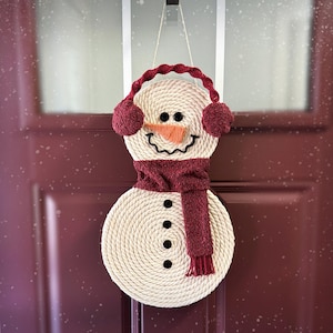 May include: A white rope snowman door hanger with a red scarf and earmuffs. The snowman has a black button nose and black eyes.