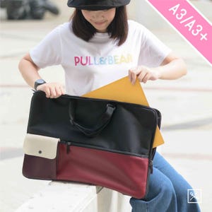 May include: A black and burgundy canvas portfolio bag with a zippered pocket and a white pocket with a button closure. The bag is large enough to hold A3 or A3+ sized paper. The bag is being held by a person wearing a white t-shirt with the text "PULL&BEAR" printed on it.