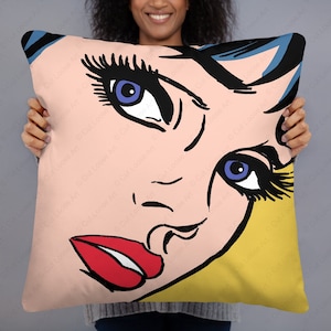 Puede incluir: Una almohada con una ilustración de estilo pop art de la cara de una mujer con ojos azules, labios rojos y pestañas negras. El fondo es amarillo y rosa.