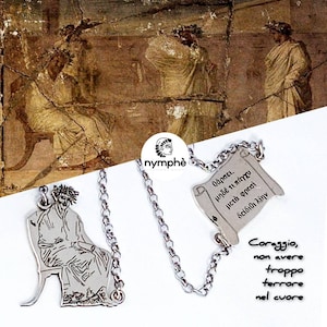 Peut inclure: Un collier argenté avec deux pendentifs : l'un représentant une figure assise, l'autre un parchemin avec du texte grec. La chaîne relie les pendentifs. En arrière-plan, une fresque antique.