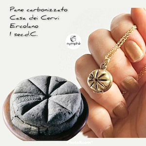 Pode incluir: Um close-up de um pão redondo, preto e carbonizado com um desenho em forma de estrela no topo. O pão está sobre um prato de madeira. Também é mostrado um colar de ouro com um pingente redondo. O pingente tem um desenho em forma de estrela. O texto "Pane carbonizzato Casa dei Cervi Ercolano 1 sec.d.C.  nymphé" é visível na imagem.