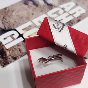 May include: A silver ring in the shape of a shovel, in a red box with a white ribbon. The ribbon has a logo with a stylized image of a person's head.
