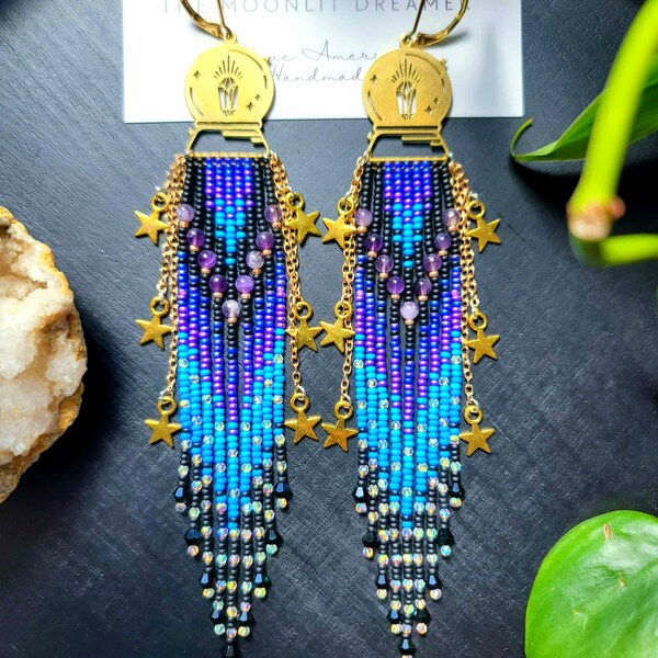May include: A pair of handmade beaded earrings with a celestial theme. The earrings feature a gold-colored top with a sunburst design, cascading blue, purple, and black beads, and gold star charms. The text "MOONLIT DREAMERS" is visible.