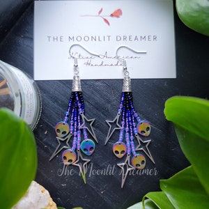 May include: A pair of iridescent alien face and star charm earrings with blue and purple beads. The earrings are handmade and feature a silver hook.