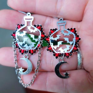 Könnte beinhalten: Silberne Ohrringe mit einem flaschenförmigen Design mit ausgeschnittenen Fledermäusen. Die Ohrringe haben einen rot-schwarzen Perlenrand, einen Mond- und Sternanhänger und eine Kette. Die Ohrringe sind für durchbohrte Ohren konzipiert.