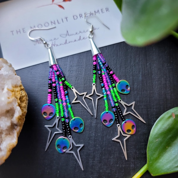 May include: A pair of handmade dangle earrings with a silver-tone metal star and alien head design. The earrings feature colorful beaded strands in pink, green, black, and blue. The text "THE MOONLIT DREAMER" is visible on a card in the background.